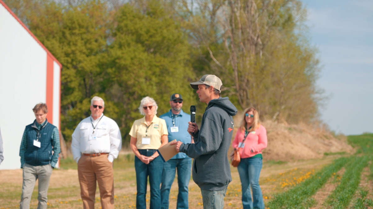 2025 Ryan Larson Man speaks to group gathered at the edge of a field.